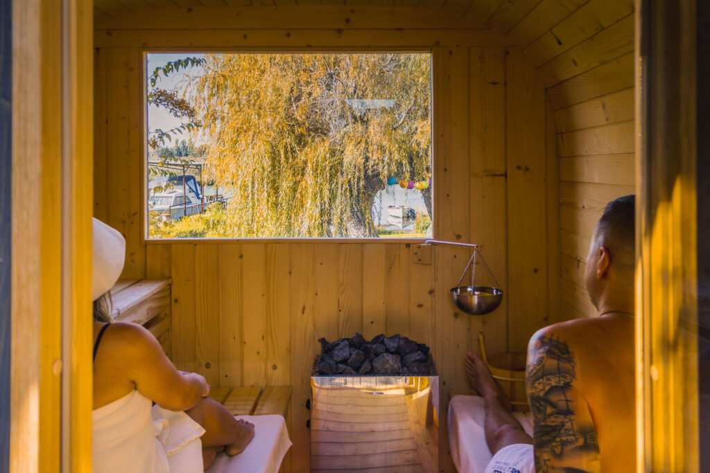 Két ember relaxál egy fa saunában, háttérben fák és hajók láthatók.